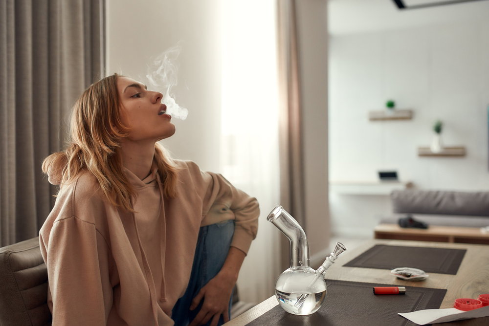 Young caucasian woman exhaling the smoke while smoking marijuana from a bong or glass water pipe, sitting in the kitchen